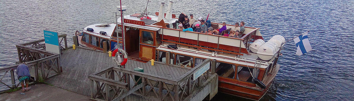 The vessel "Tuulispää" at a wooden pier with water and green forest in the background.